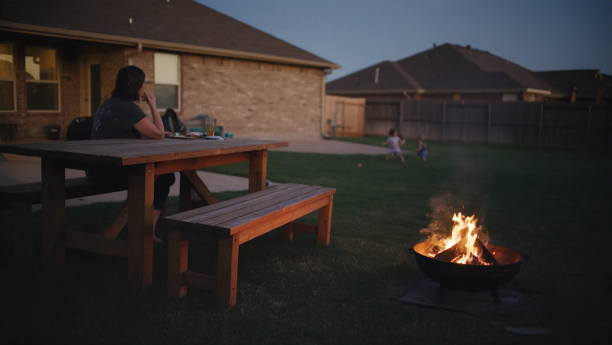 A backyard at dusk: a woman at a cedar picnic table with her back to us, a cedar bench, a fire in a steel pit, two children playing far off on the lawn